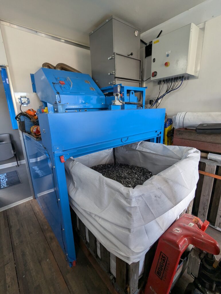 Industrial blue shredding machine used by BCC IT to destroy data drives safely, feeding fragments into a collection bag for recycling.