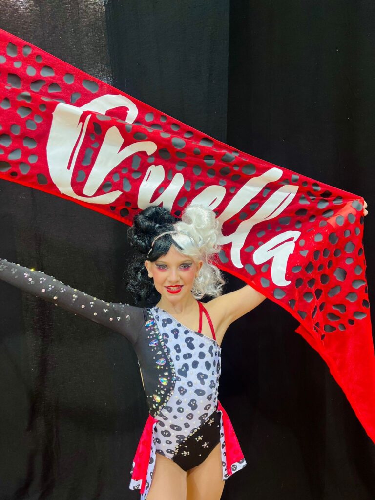 Chloe performing as Cruella Deville for her routine in black-and-white-costume holding a red Cruella flag backdrop