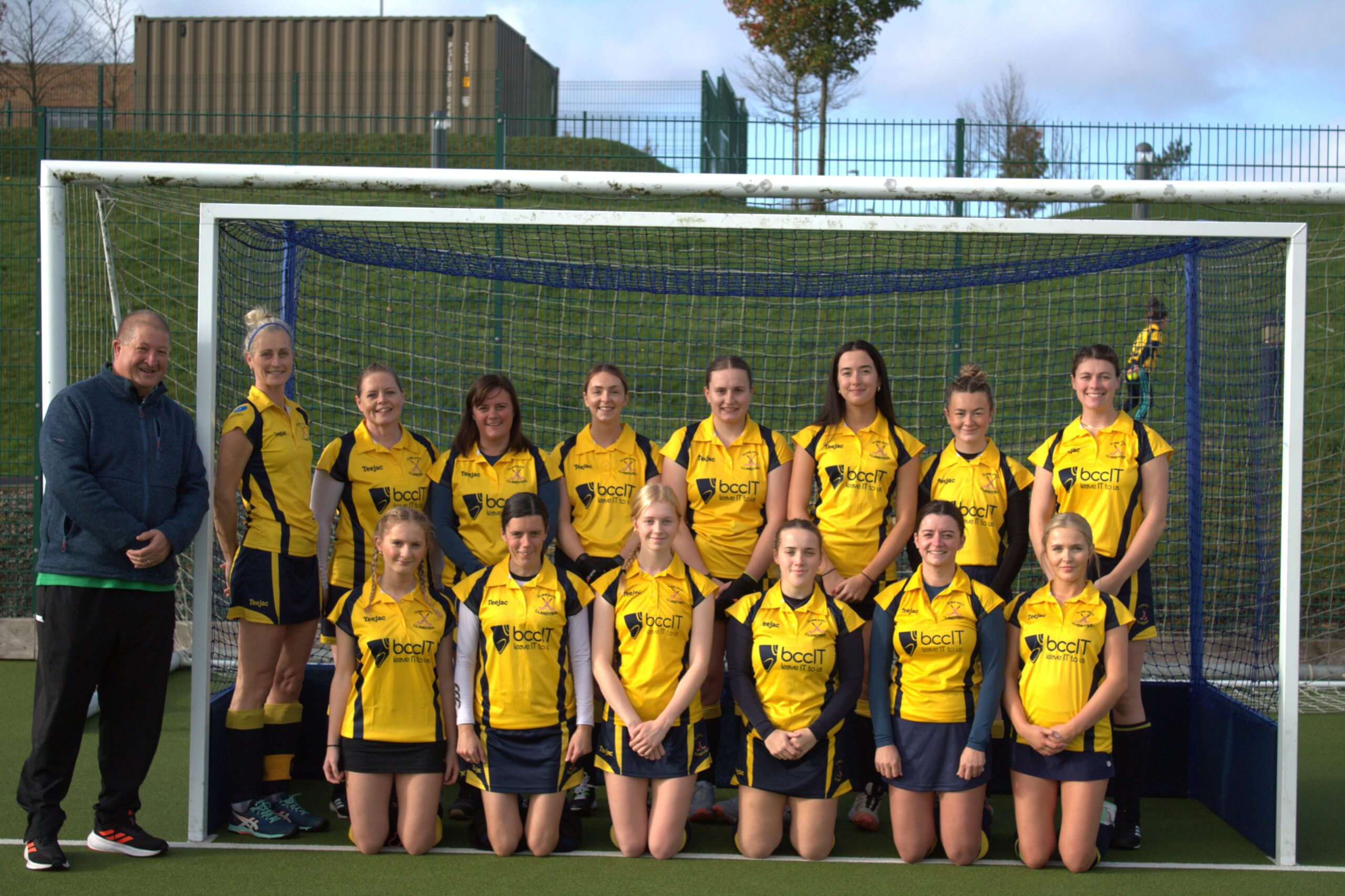 BCC IT Director Hywel Ifans standing with the Clwb Hoci Llandysul senior girls' team in yellow kits displaying the bccit logo in front of the goal on the Ysgol Bro Teifi astro pitch