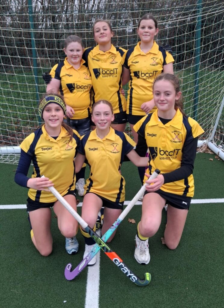 Clwb Hoci Llandysul junior girls' hockey team in yellow kits displaying the bccit logo in front of the goal on the Ysgol Bro Teifi astro pitch posing with crossed hockey sticks.